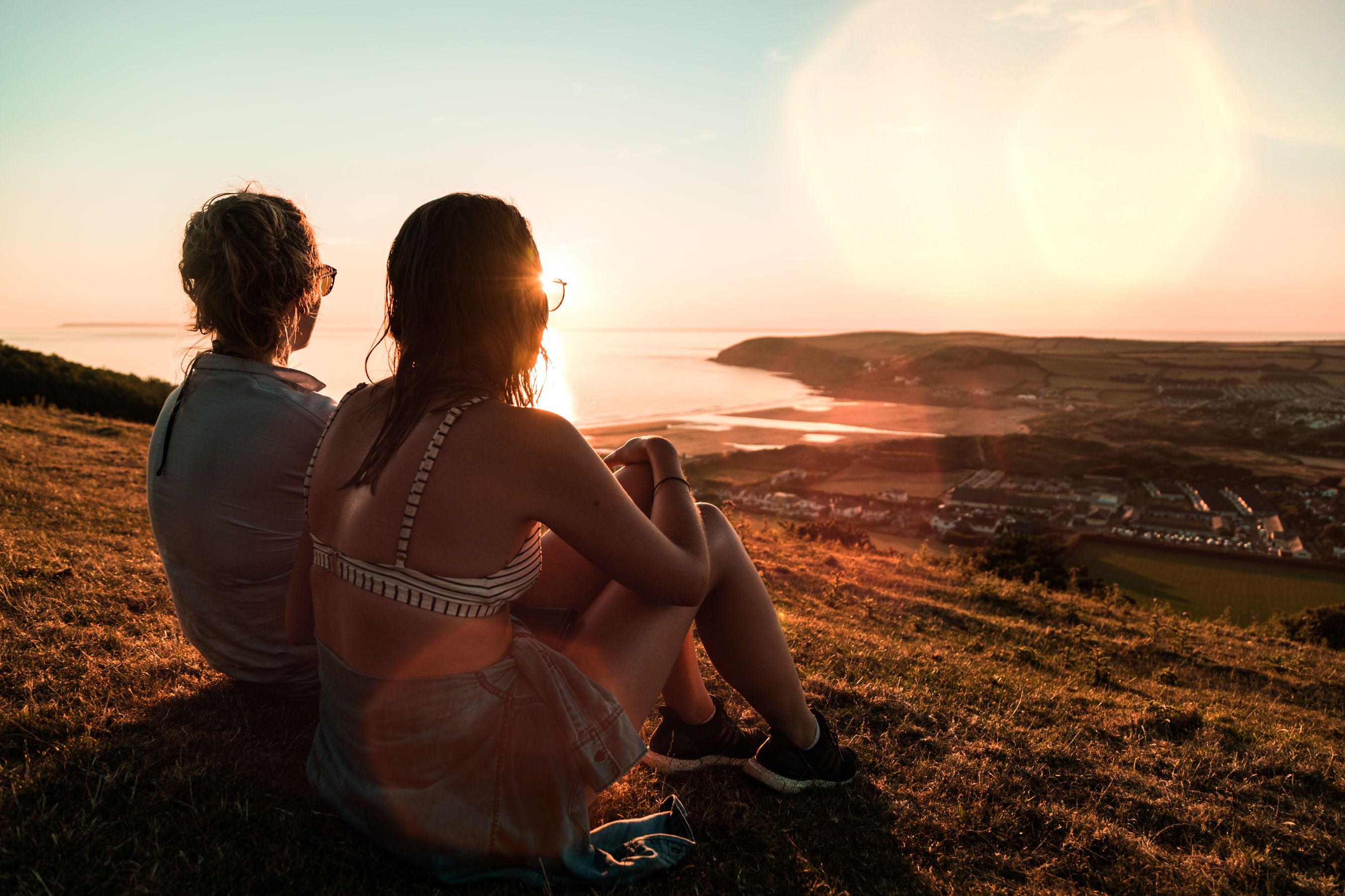 Two girls sitting on hillside looking over seaside village. 